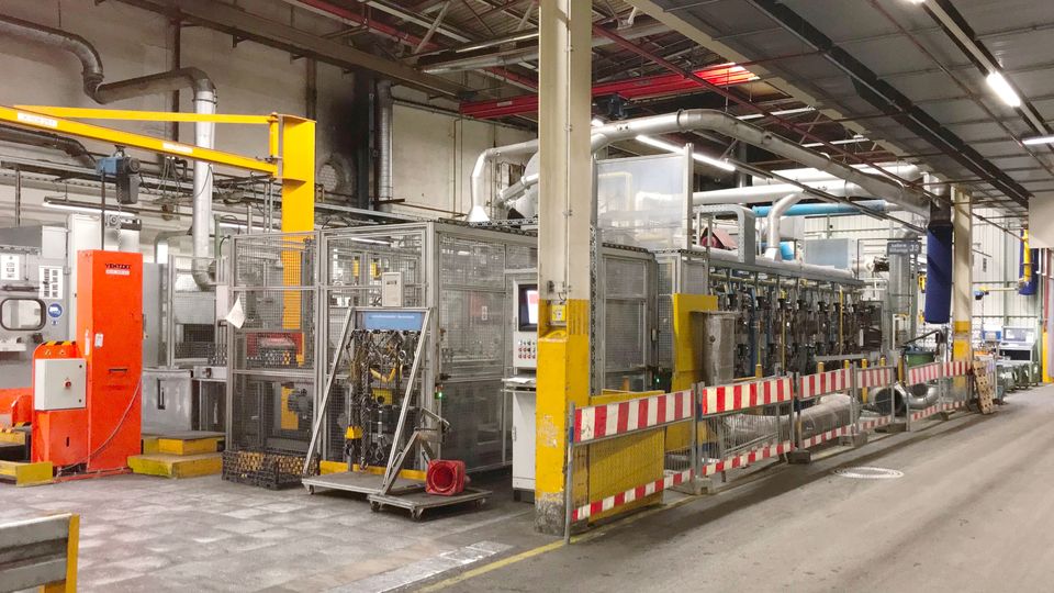 Factory interior with machinery and safety barriers. Industrial equipment and pipes are visible under a high ceiling, with concrete flooring.