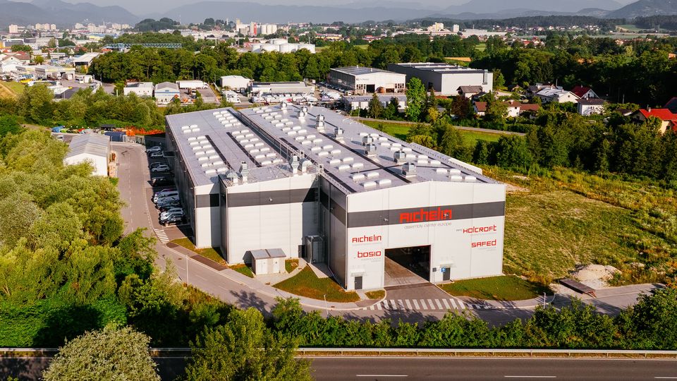 Aerial view of a large industrial warehouse surrounded by greenery, with mountains in the background and a road in the foreground.