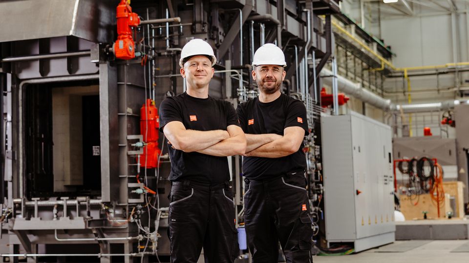 Two workers in helmets and black uniforms stand confidently in front of industrial machinery inside a spacious factory.