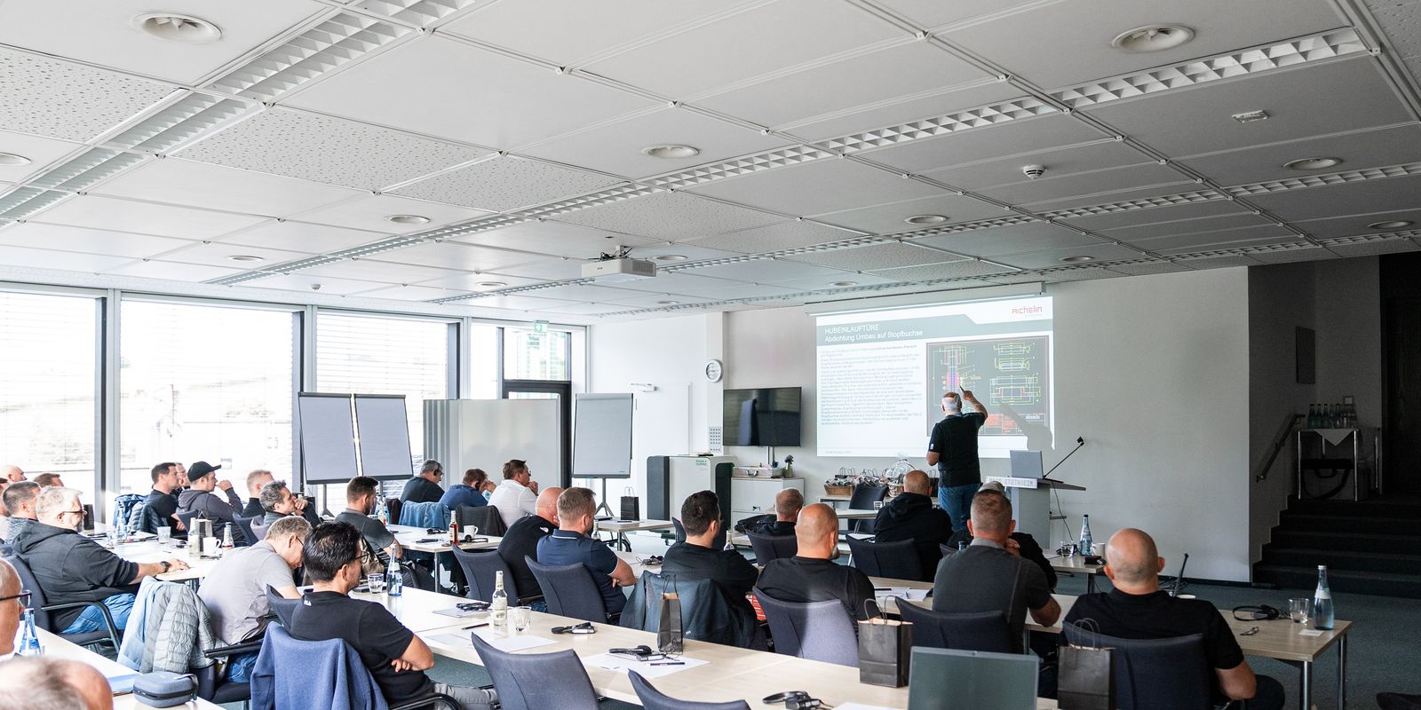 A group of people in a classroom setting listening to a presentation with slides projected on a screen. Laptops and notepads are on tables.