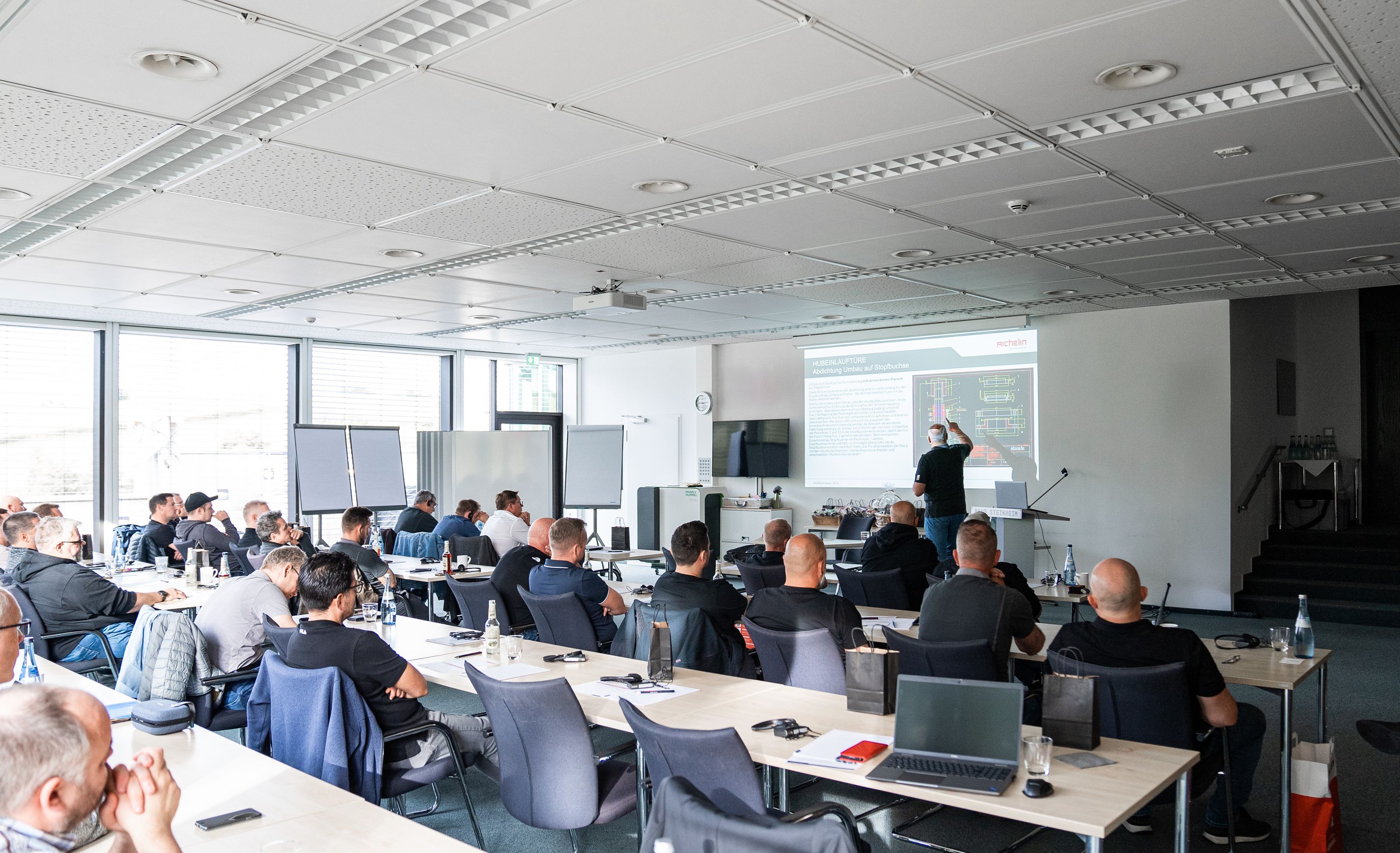 A group of people in a classroom setting listening to a presentation with slides projected on a screen. Laptops and notepads are on tables.