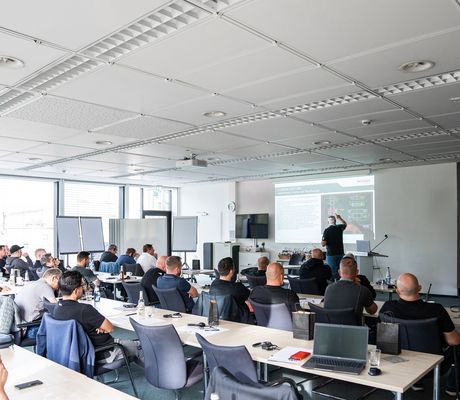 A group of people in a classroom setting listening to a presentation with slides projected on a screen. Laptops and notepads are on tables.