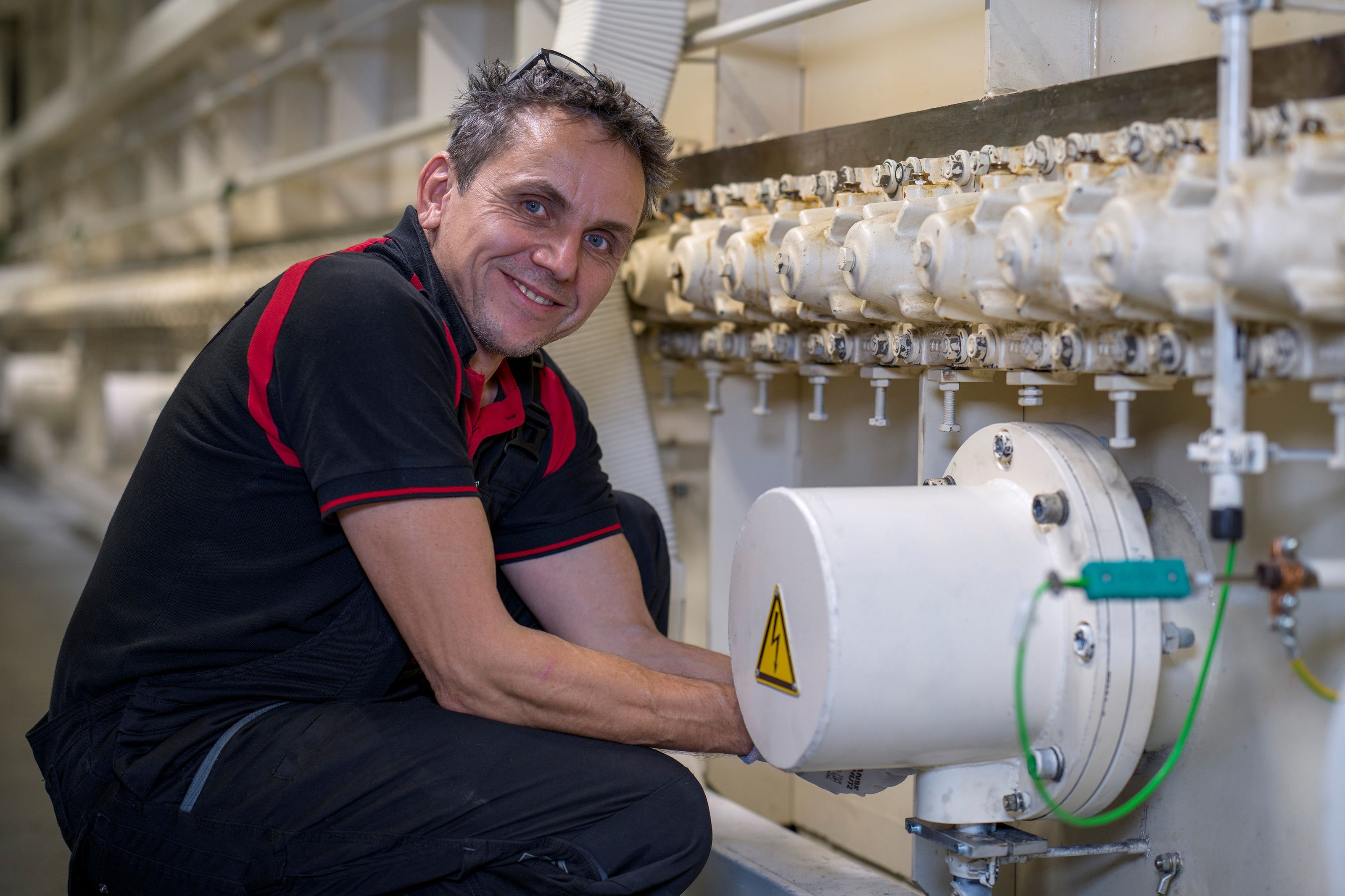 Man in a black and red shirt smiles while working on industrial machinery, adjusting a pipe with multiple valves.