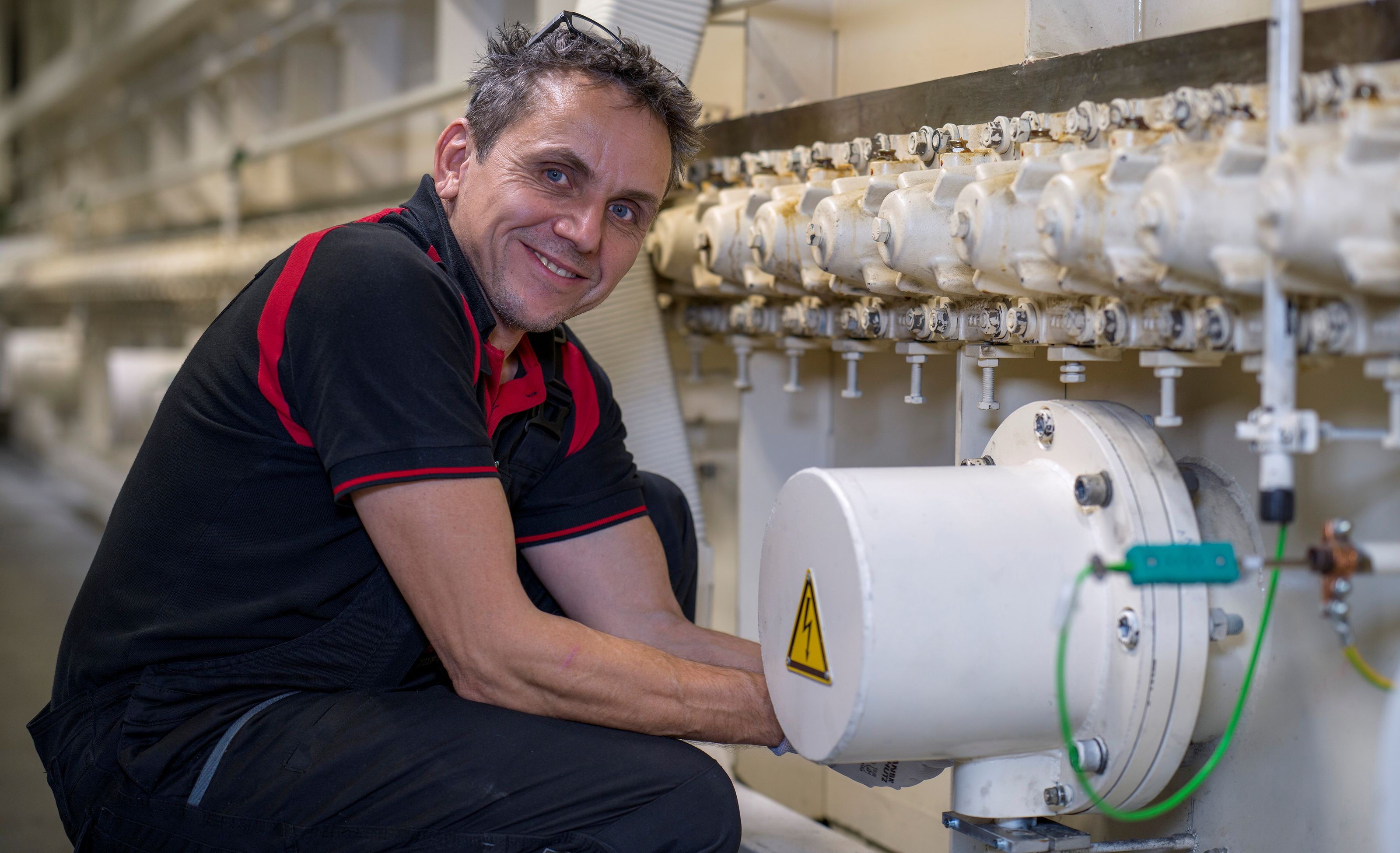 Man in a black and red shirt smiles while working on industrial machinery, adjusting a pipe with multiple valves.