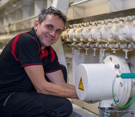 Man in a black and red shirt smiles while working on industrial machinery, adjusting a pipe with multiple valves.