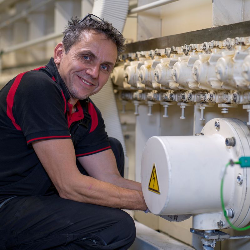 Man in a black and red shirt smiles while working on industrial machinery, adjusting a pipe with multiple valves.