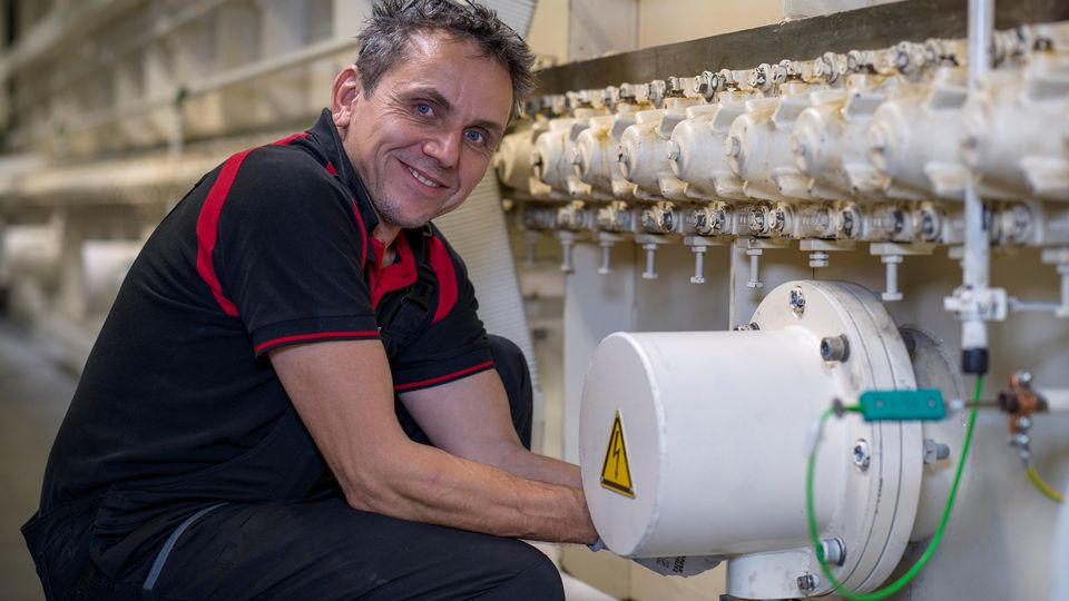 Man in a black and red shirt smiles while working on industrial machinery, adjusting a pipe with multiple valves.