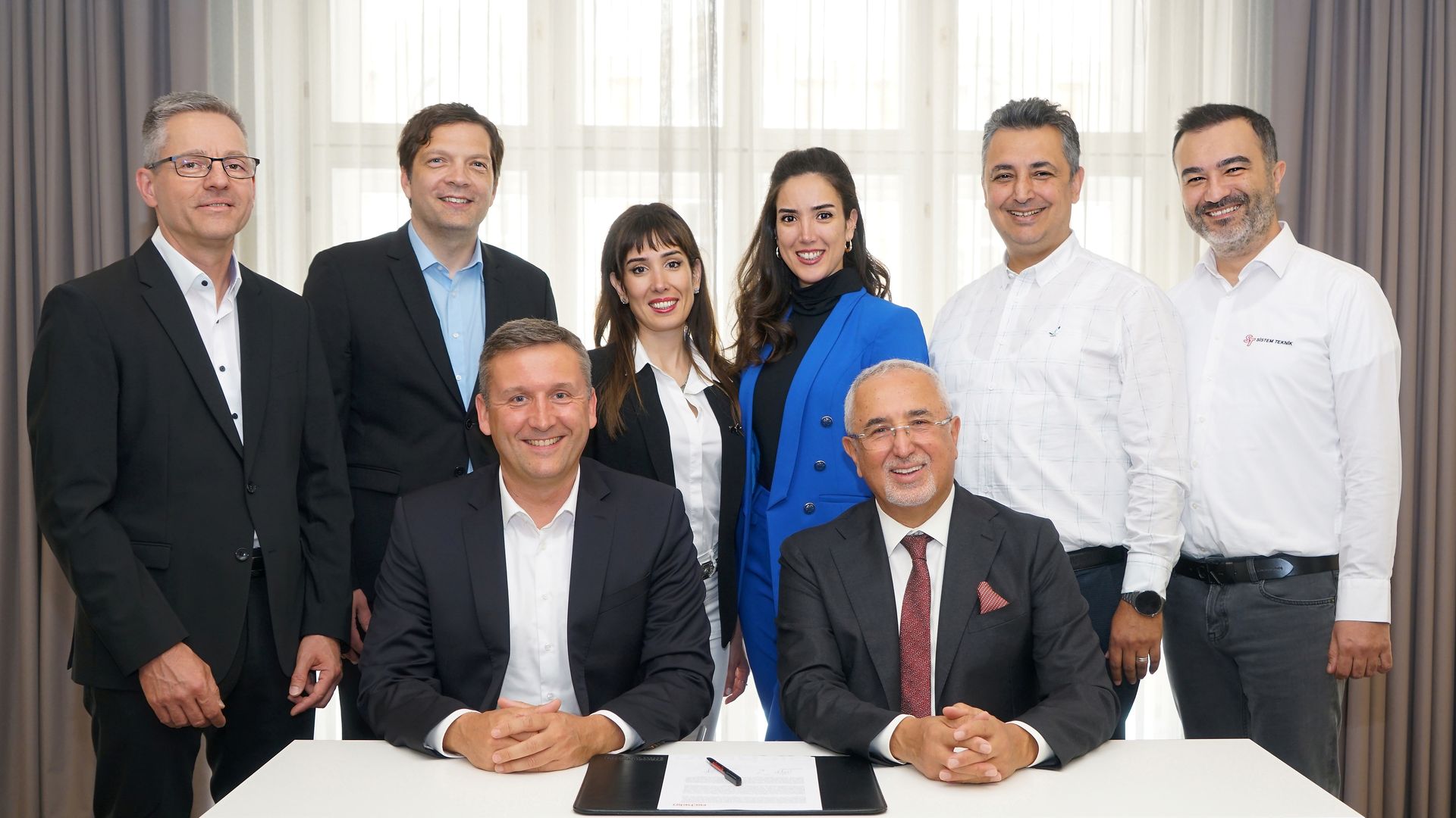 Eight people in business attire, six standing and two seated at a table with a document, posing for a group photo indoors.