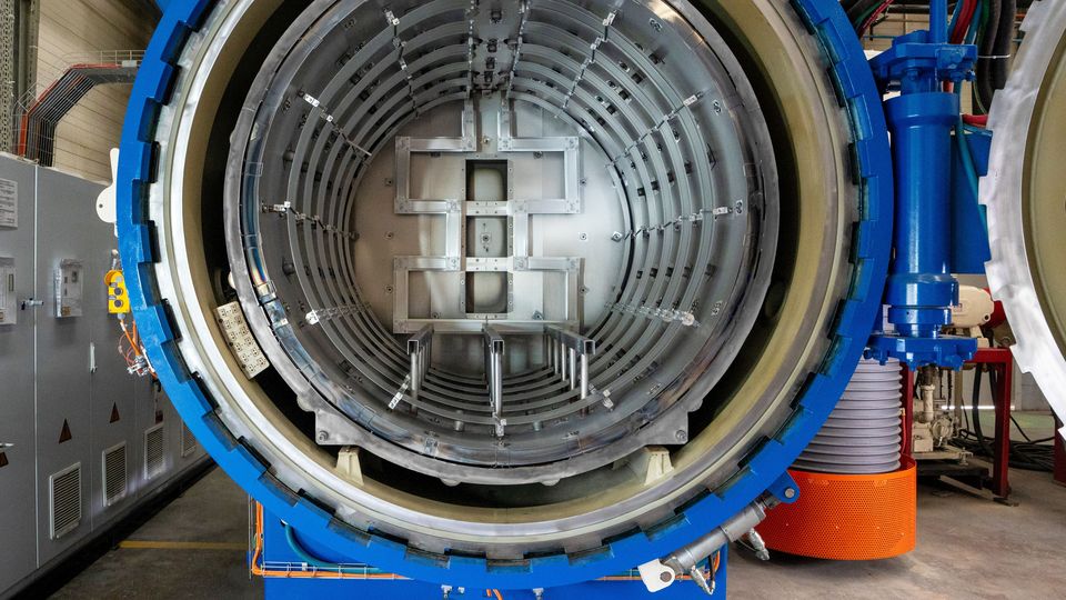 Open industrial autoclave with metal interior and blue exterior, situated in a factory setting with visible machinery and equipment.