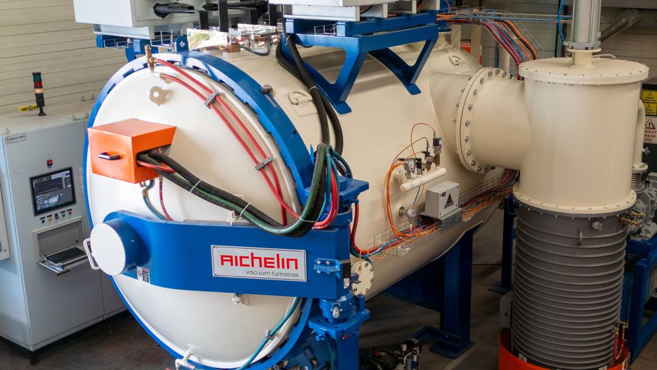 Industrial vacuum furnace with control panel, wires, and gauges in a workshop setting. The furnace is large and white with blue and orange components.