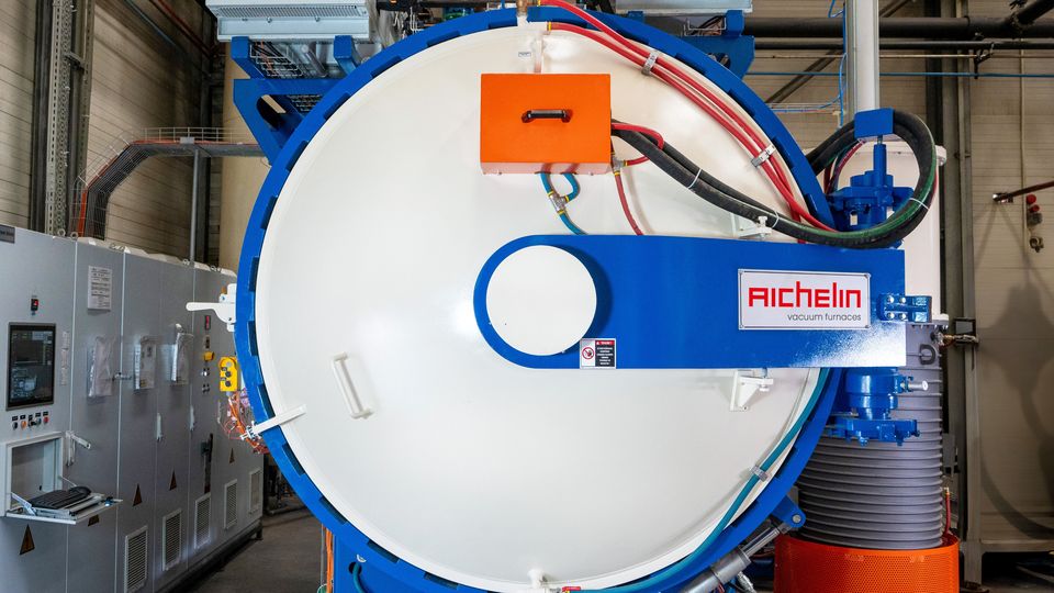 Large industrial vacuum furnace with blue and orange components, control panels, and cables in a factory setting.