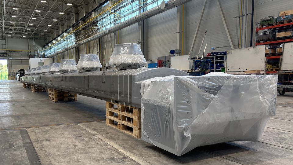 A long, industrial metal component covered in protective plastic sits on wooden pallets inside a spacious warehouse with high ceilings.