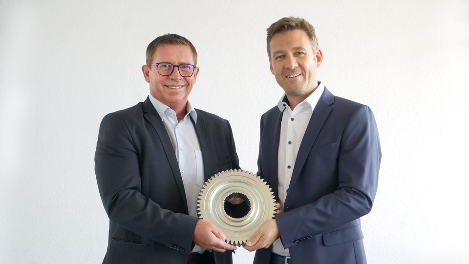 Two men in suits hold a large, metallic gear together, smiling against a plain white background.