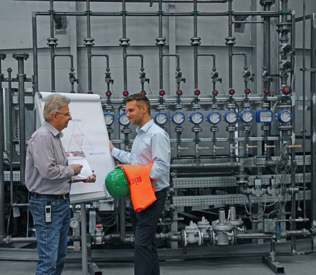Two men discussing by a flip chart in an industrial setting, surrounded by pipes and gauges, with a green helmet and orange safety vest nearby.