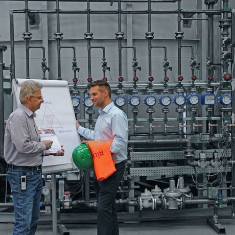 Two men discussing by a flip chart in an industrial setting, surrounded by pipes and gauges, with a green helmet and orange safety vest nearby.