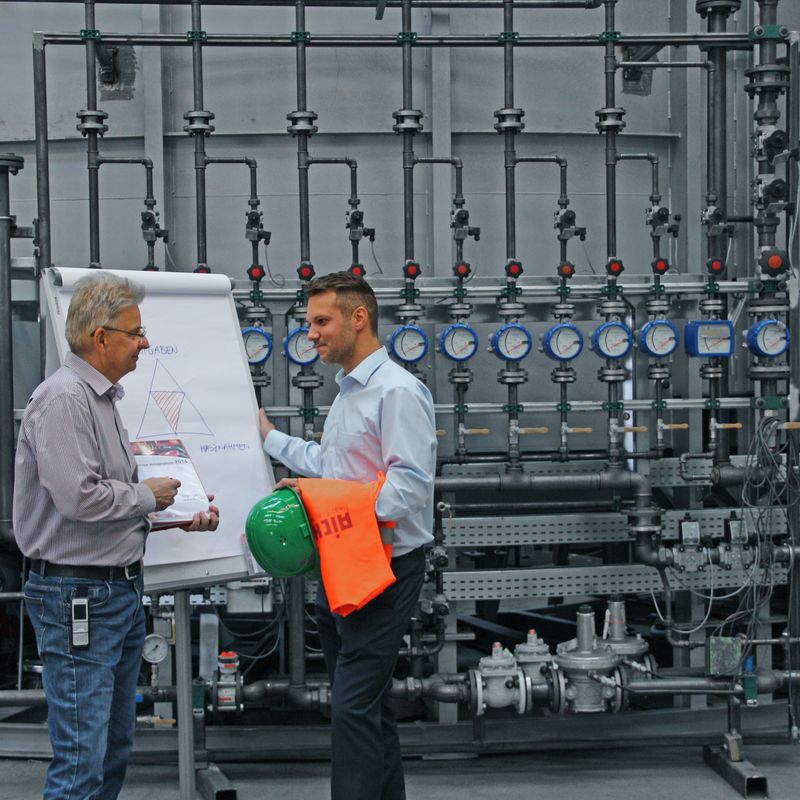 Two men discussing by a flip chart in an industrial setting, surrounded by pipes and gauges, with a green helmet and orange safety vest nearby.