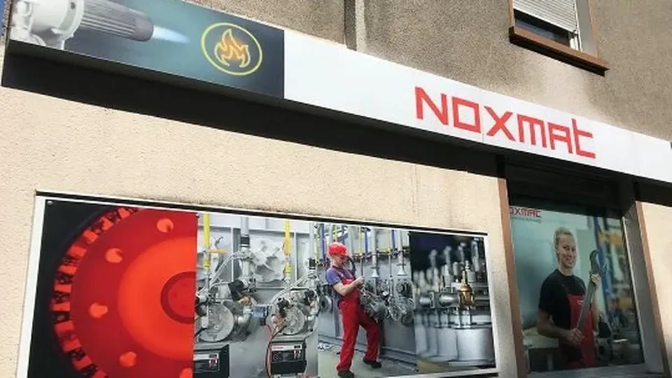 Ladenfront mit „NOXMAT”-Beschriftung. Zeigt Bilder von Industrieanlagen und einem Arbeiter in roter Uniform und Schutzhelm.