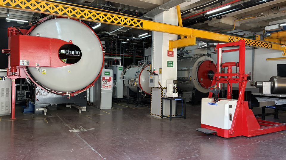 Industriehalle mit gelbem Kran, rotem Vakuumofen und Ausrüstungen. Betonboden und technische Installationen an der Decke.