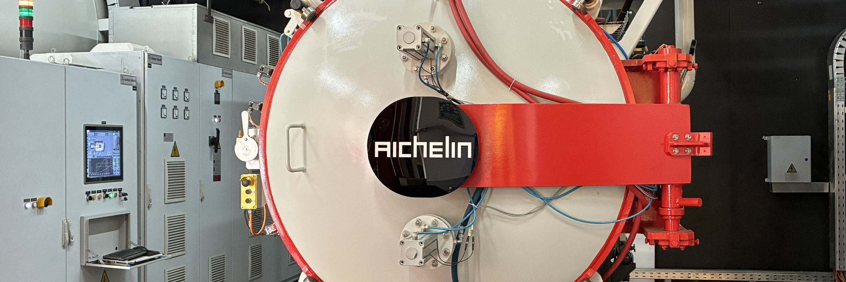 Industrieller Vacuumofen von Aichelin in einer Fabrikumgebung, umgeben von Rohrleitungen und Kontrollsystemen.