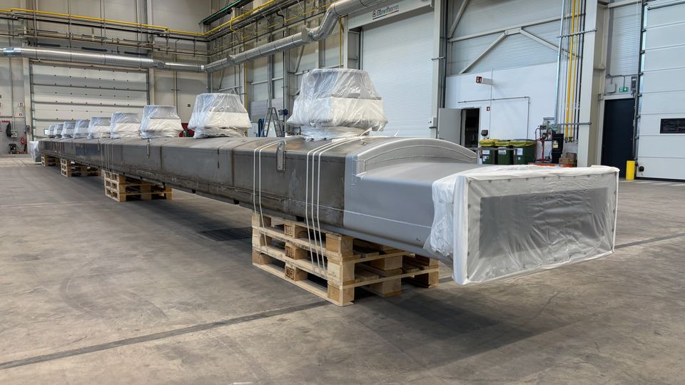 A long industrial component wrapped in protective material rests on wooden pallets inside a spacious warehouse, surrounded by machinery and equipment.