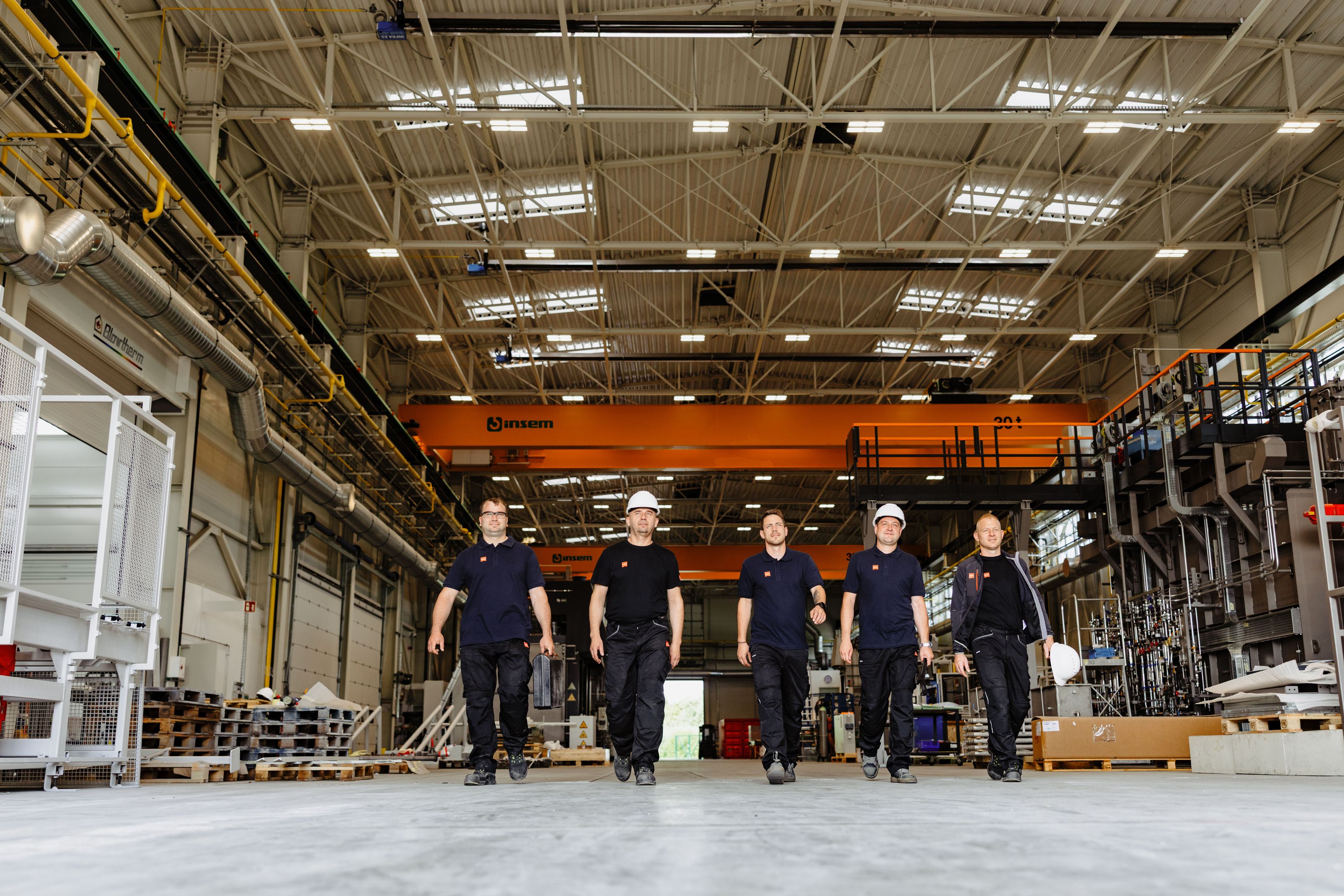 Five workers in safety gear walk confidently through a spacious, well-lit industrial facility, with machinery and equipment in the background.