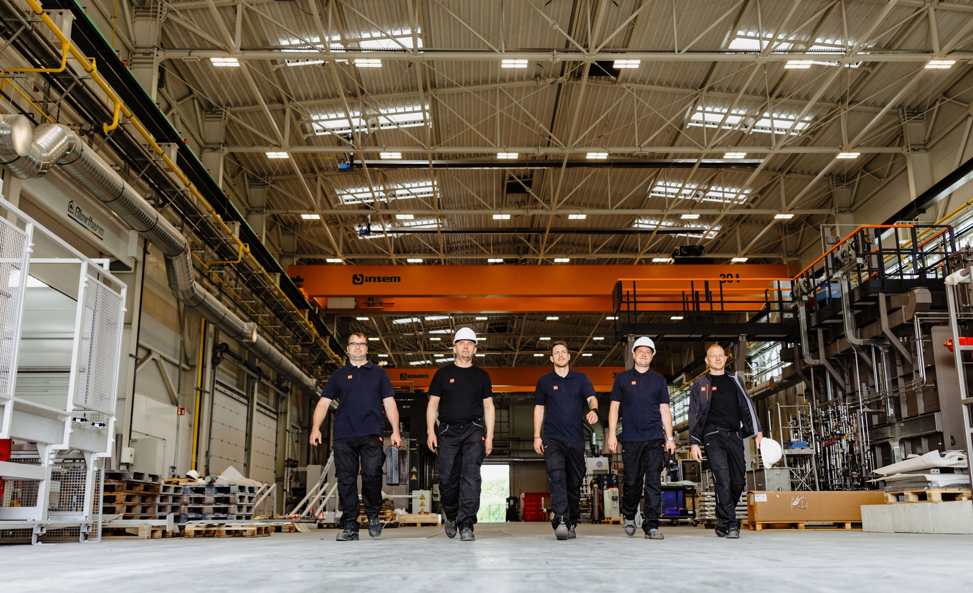 Five workers in safety gear walk confidently through a spacious, well-lit industrial facility, with machinery and equipment in the background.