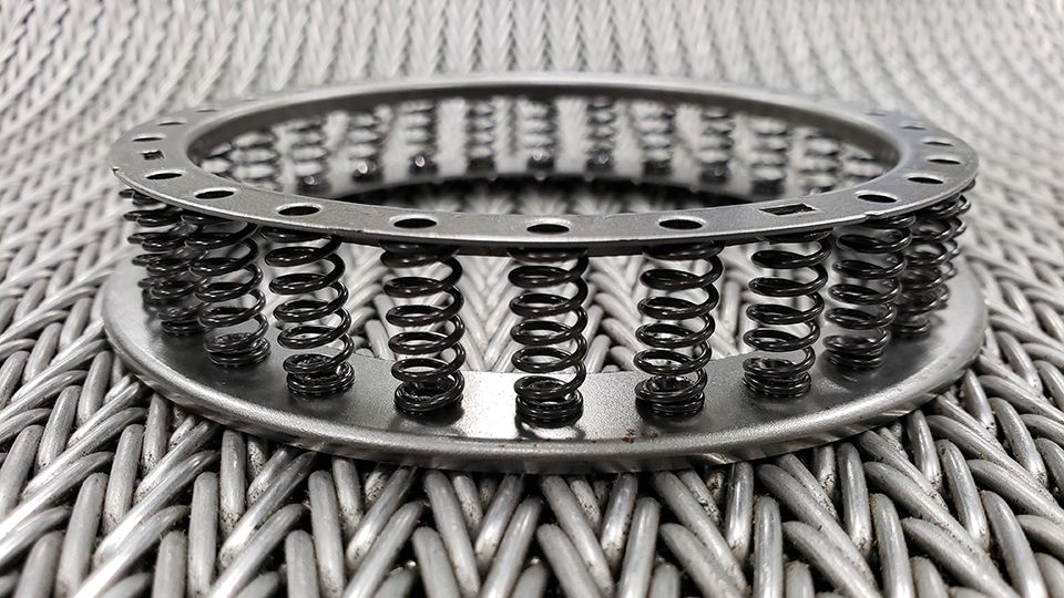 A round metal component with multiple small springs between two perforated circular plates, placed on a textured surface.