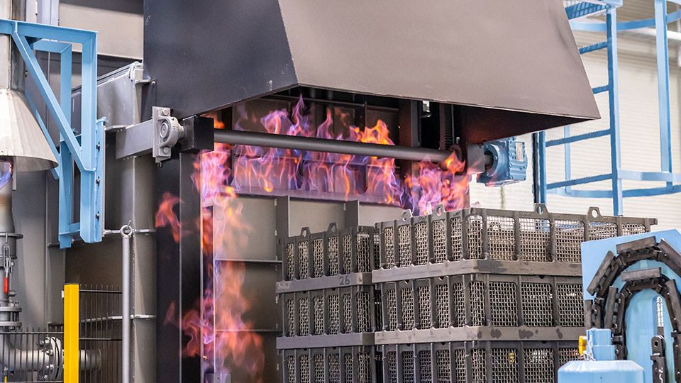 Industrial furnace with bright orange flames processing stacked metal crates inside a factory setting.