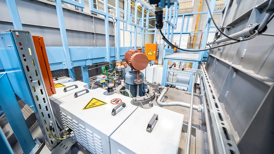 Industrial machinery with control panels, wires, and warning signs inside a metal-framed facility, featuring a large cylinder and piping system.