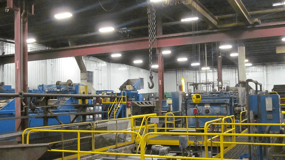 Interior of an industrial factory with machinery, yellow safety rails, and a large overhead crane with a chain and hook hanging down.