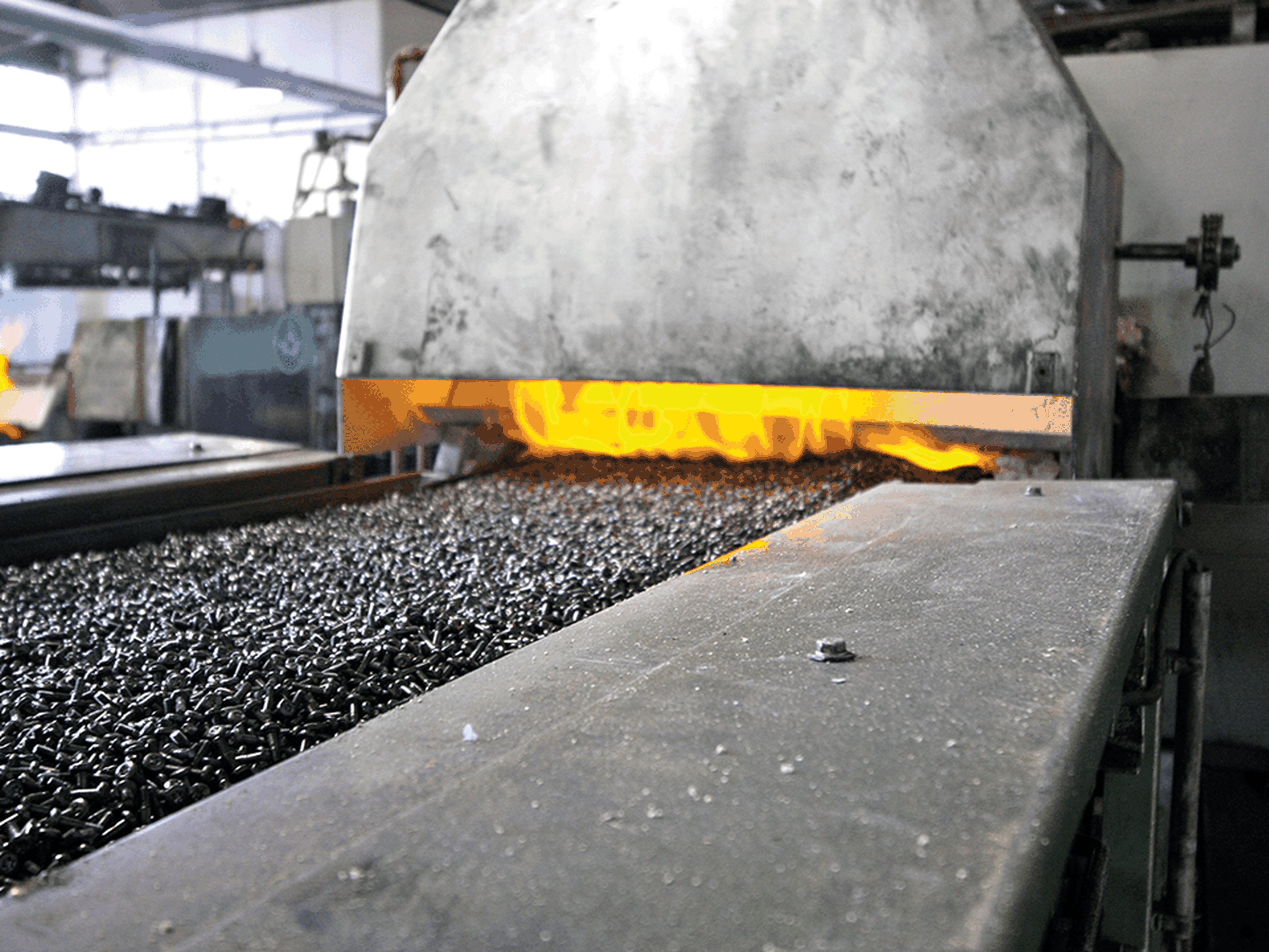 Metal parts on a conveyor belt enter a furnace with visible flames, indicating a high-temperature industrial process.