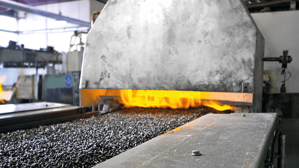Metal parts on a conveyor belt enter a furnace with visible flames, indicating a high-temperature industrial process.