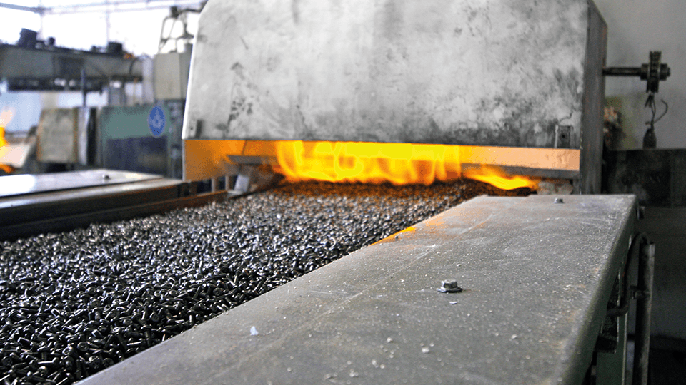 Metal parts on a conveyor belt enter a furnace with visible flames, indicating a high-temperature industrial process.