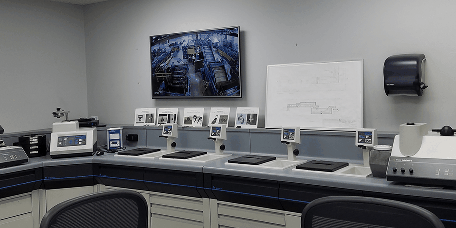 A modern laboratory room with desks, various equipment, a monitor displaying a factory floor, and a whiteboard with diagrams on the wall.