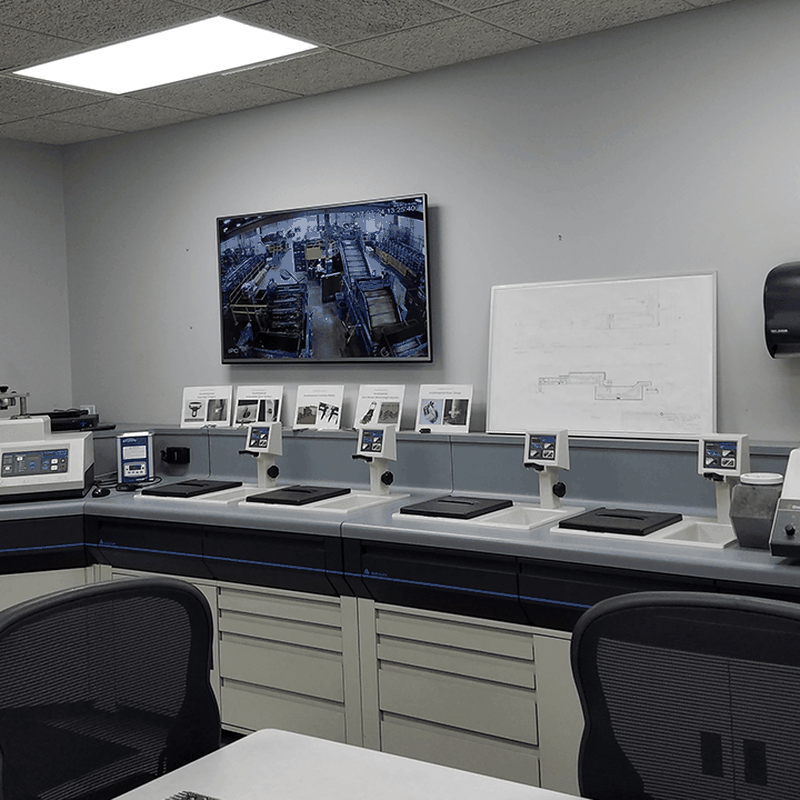 A modern laboratory room with desks, various equipment, a monitor displaying a factory floor, and a whiteboard with diagrams on the wall.