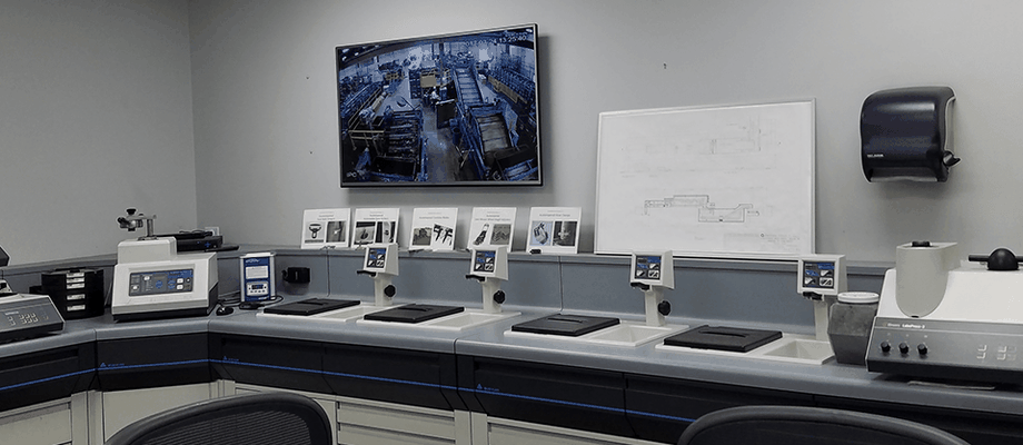 A modern laboratory room with desks, various equipment, a monitor displaying a factory floor, and a whiteboard with diagrams on the wall.