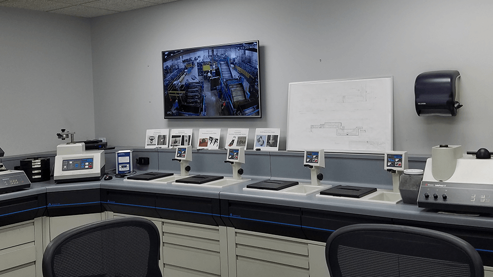 A modern laboratory room with desks, various equipment, a monitor displaying a factory floor, and a whiteboard with diagrams on the wall.