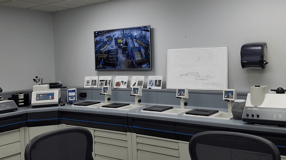 A modern laboratory room with desks, various equipment, a monitor displaying a factory floor, and a whiteboard with diagrams on the wall.