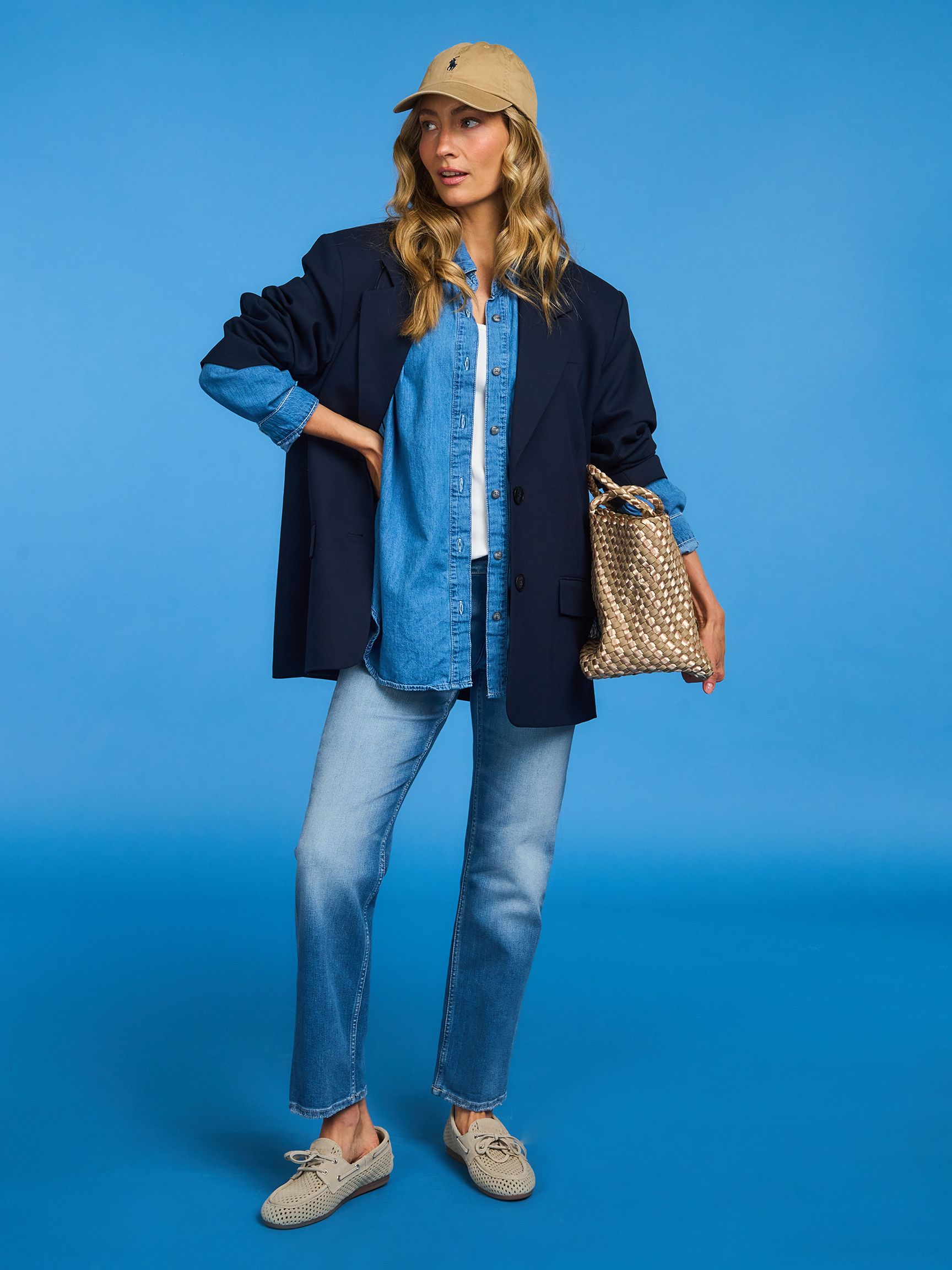 Woman in a navy blazer and denim outfit, wearing a tan cap and sneakers, holding a woven bag, stands against a blue background.