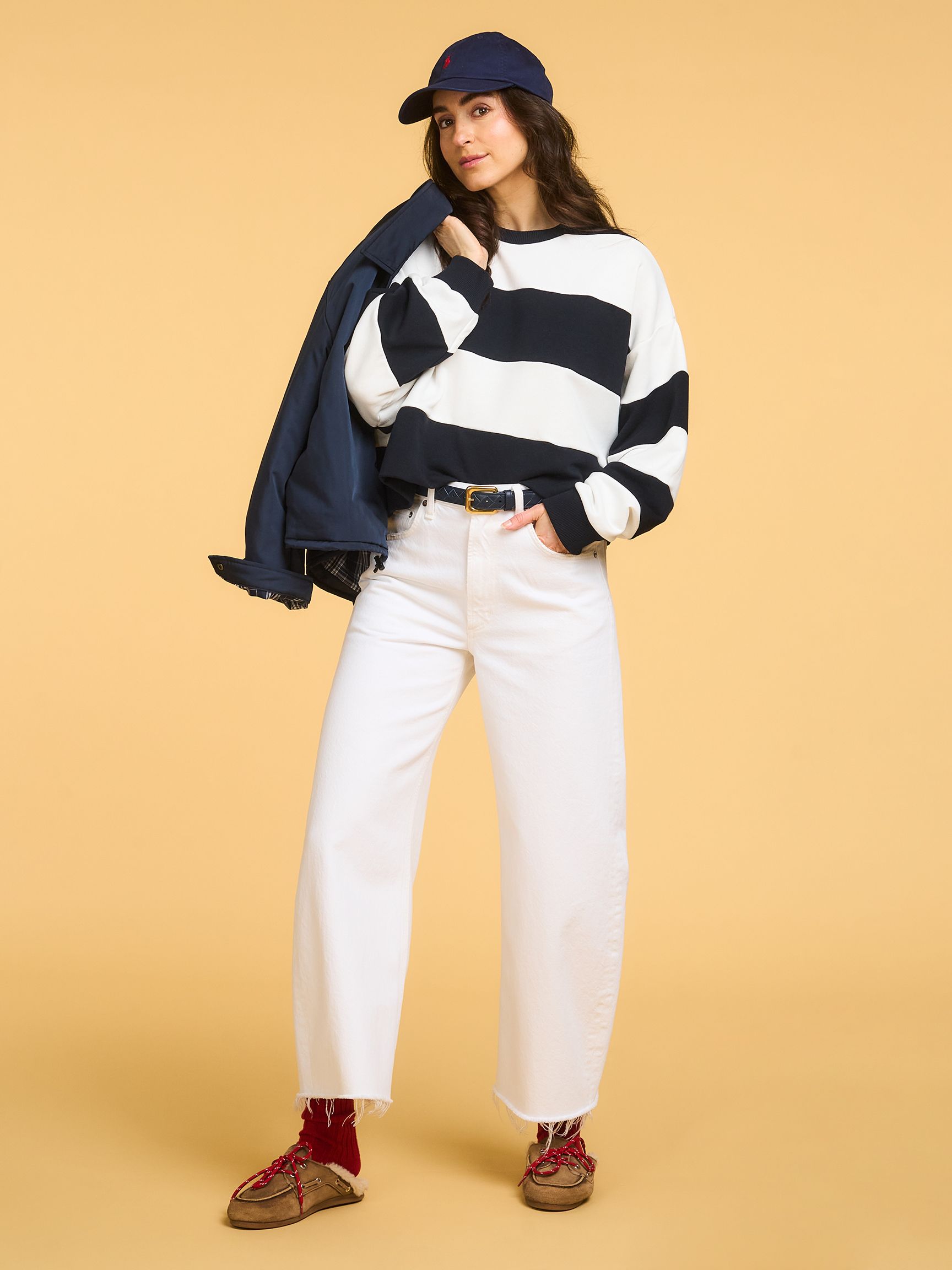 Woman in a striped sweater, white pants, red socks, and a navy cap poses casually against a beige background, holding a jacket.