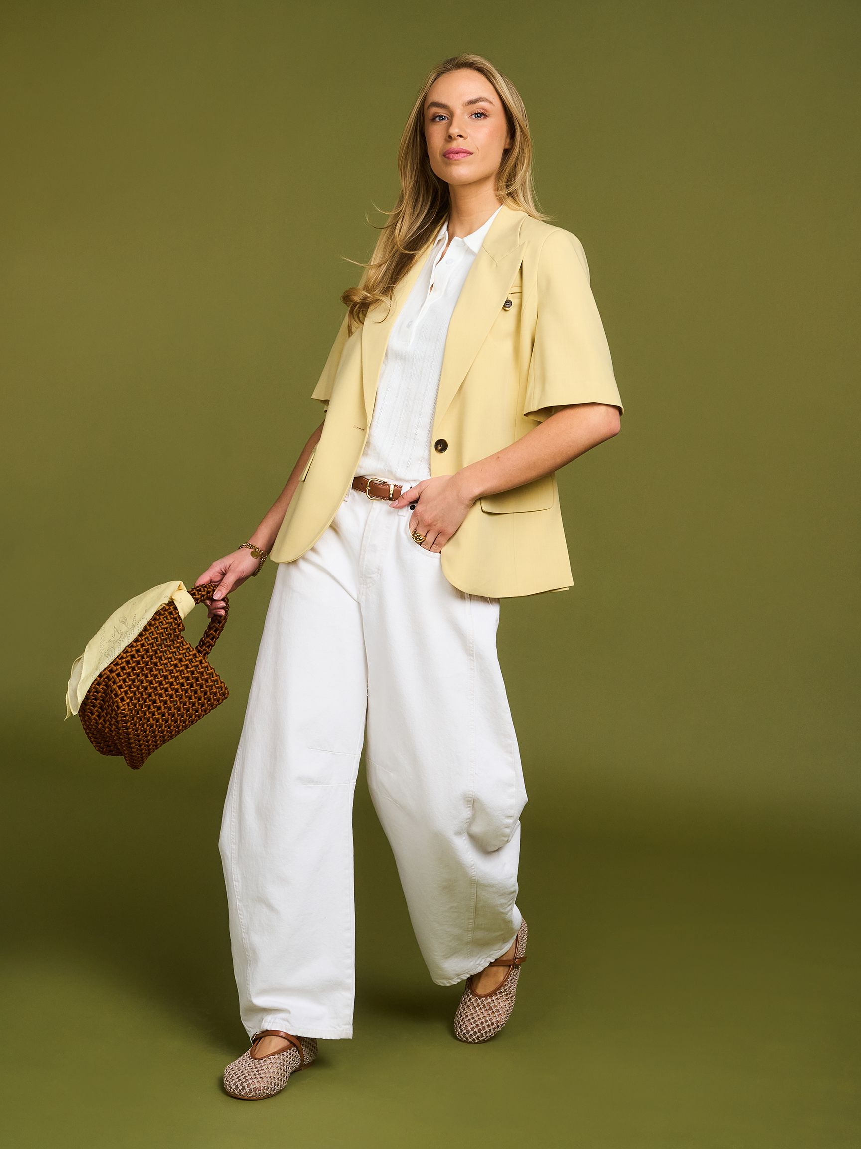 A woman in a yellow blazer and white outfit holds a brown patterned handbag, standing on a green background.