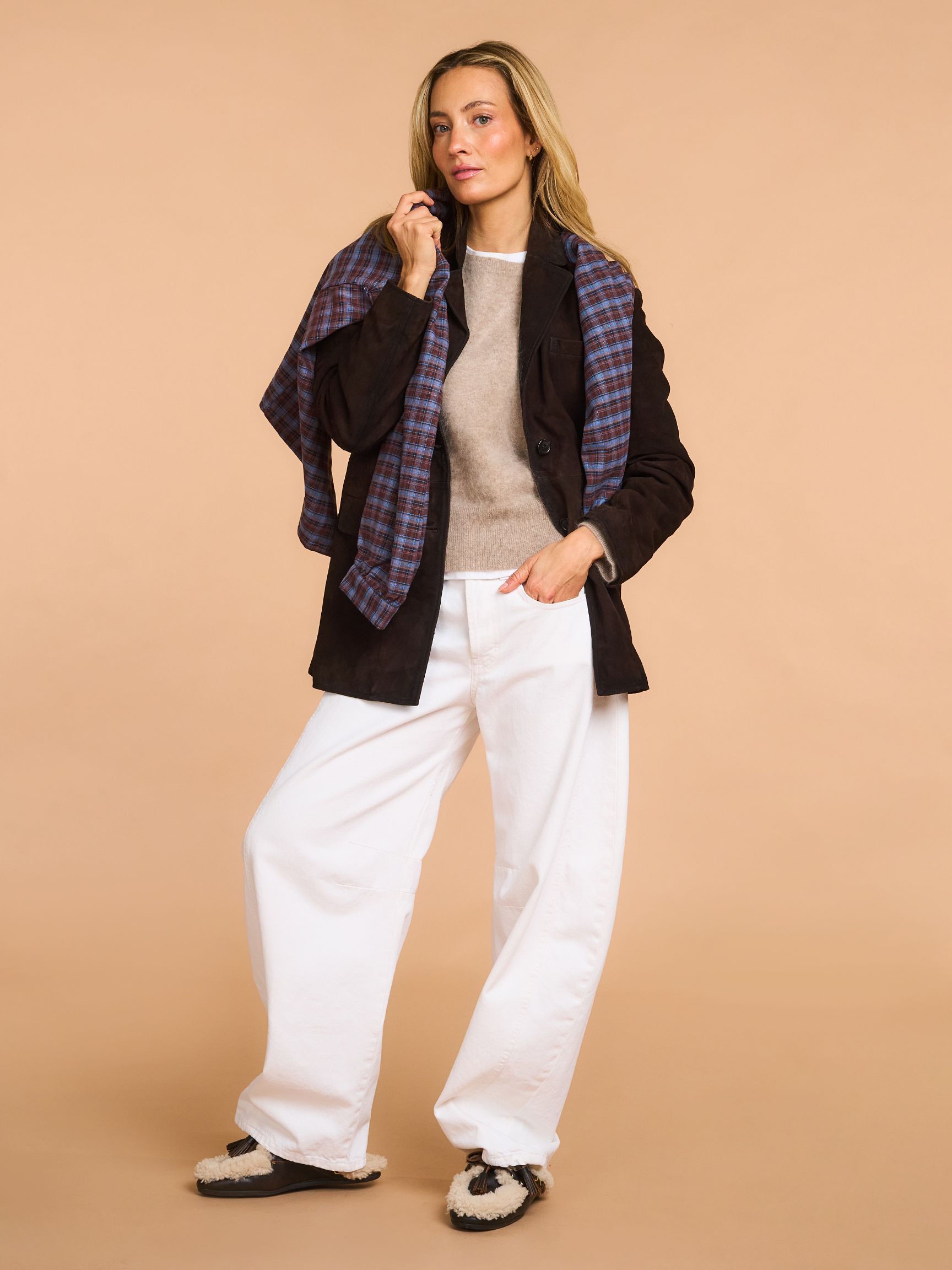 Person posing confidently in a brown jacket with a plaid scarf, beige sweater, white wide-leg pants, and black slippers on a beige background.