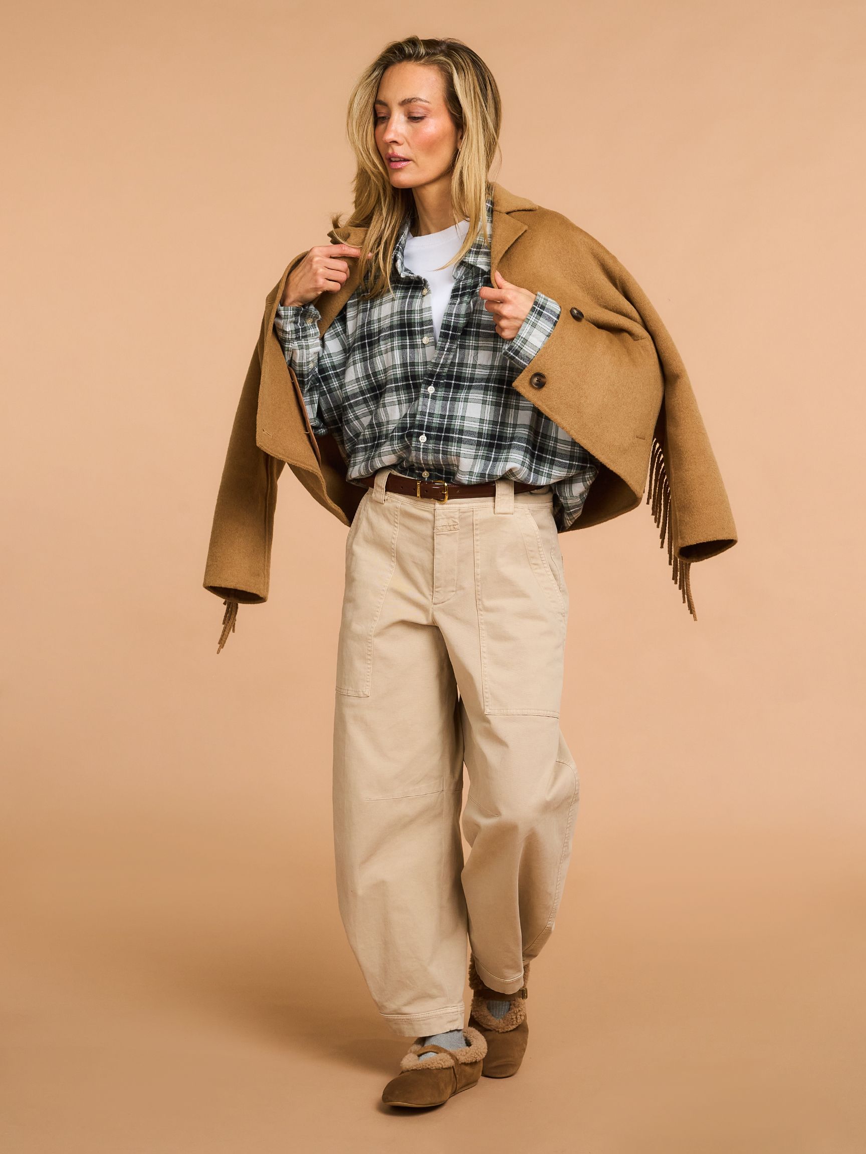 Woman in a plaid shirt and beige pants with a brown coat draped over her shoulders, standing against a tan background.