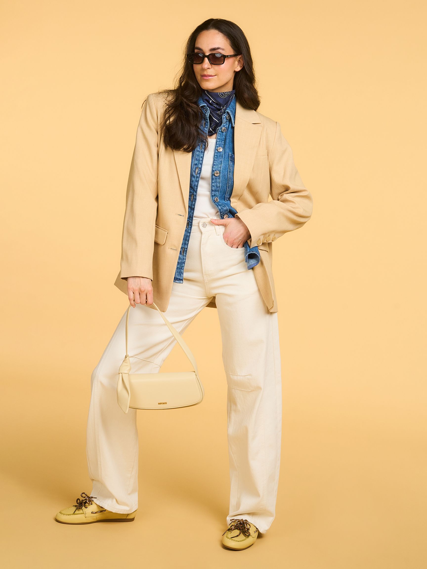 Woman in beige blazer, denim shirt, white pants, sunglasses, and bandana, posing with a beige handbag against a yellow background.