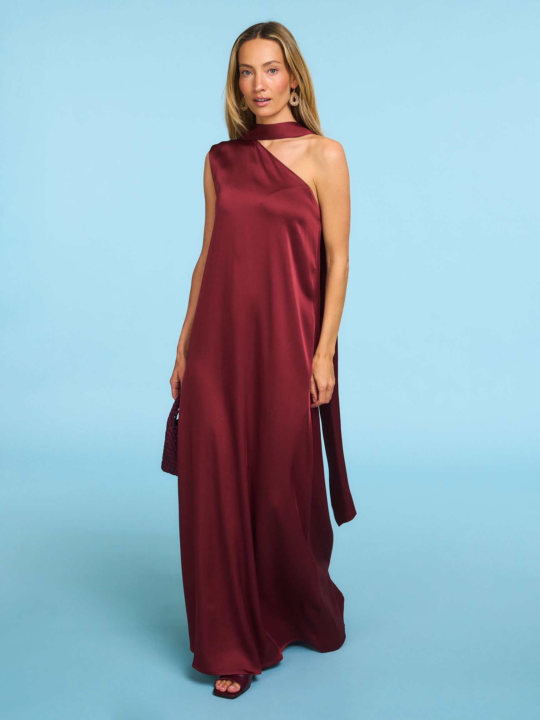 Woman in a flowing burgundy gown with a halter neck stands against a light blue background, holding a small clutch.