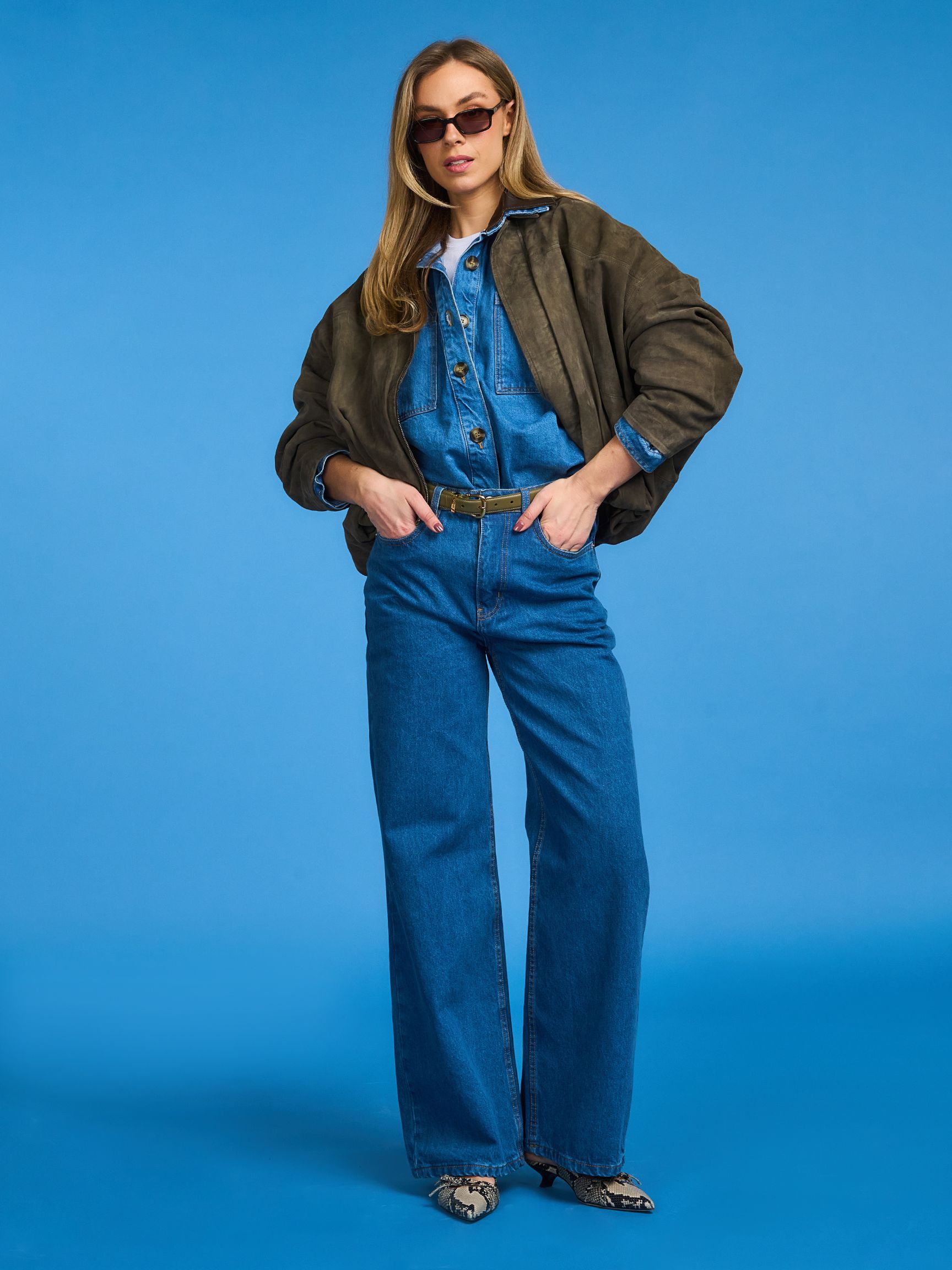 Person wearing sunglasses, a green jacket over a denim jumpsuit, and snakeskin shoes stands against a blue background.