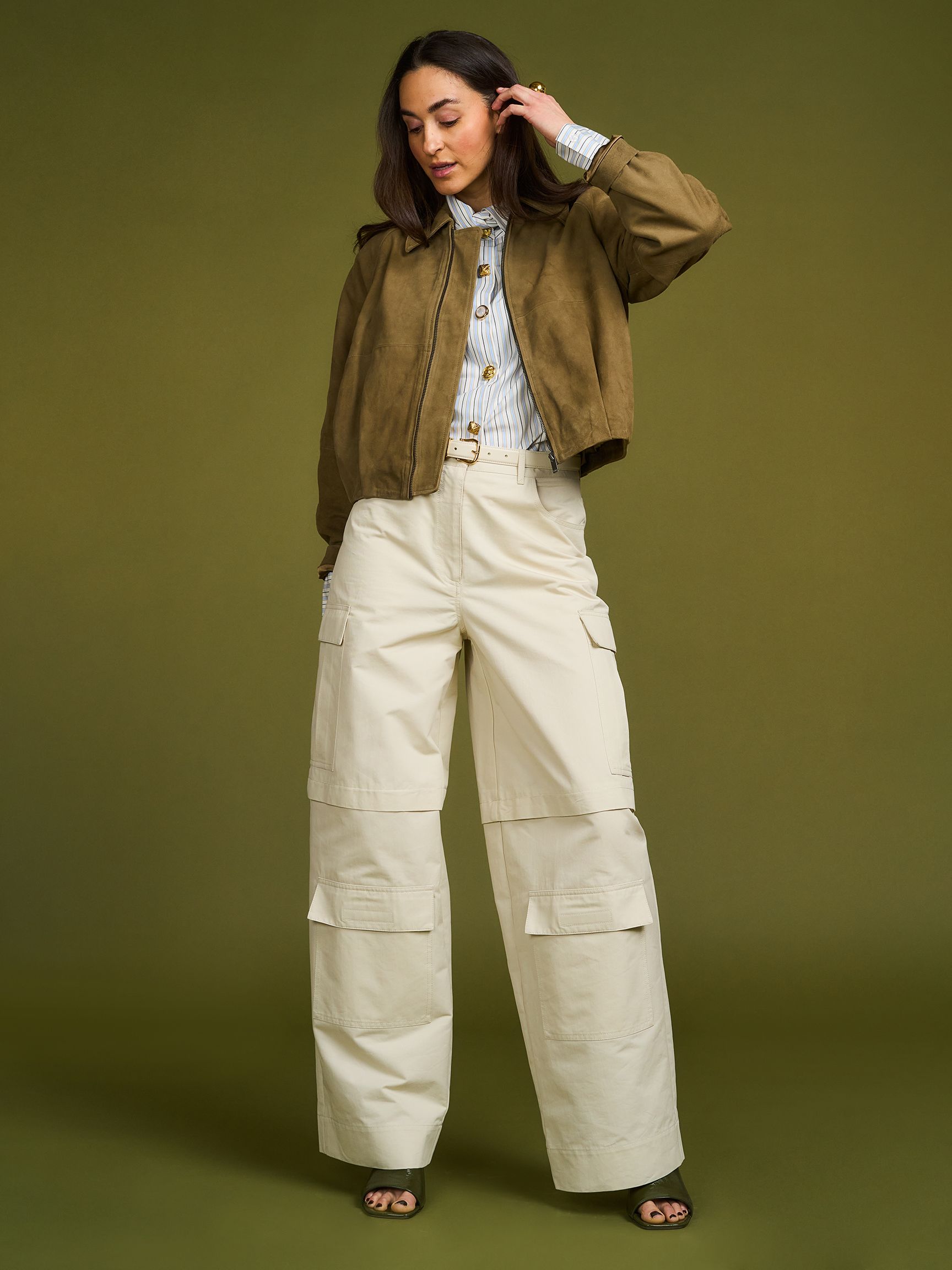 Woman in a brown jacket and white wide-leg pants stands against an olive green background, looking down with one hand raised to her hair.