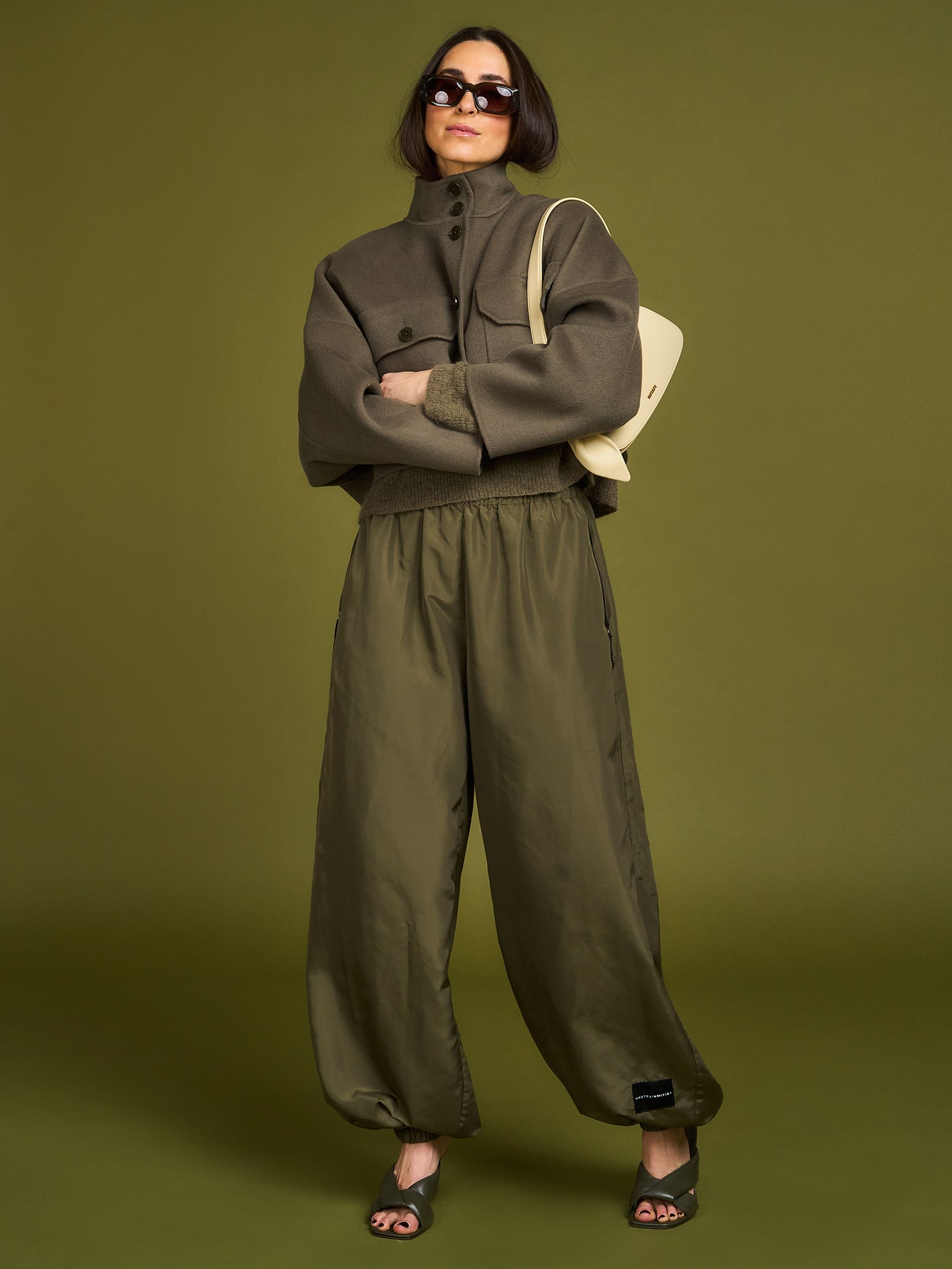 A person wearing a stylish oversized jacket, baggy pants, sunglasses, and heeled sandals, holding a bag against an olive green background.