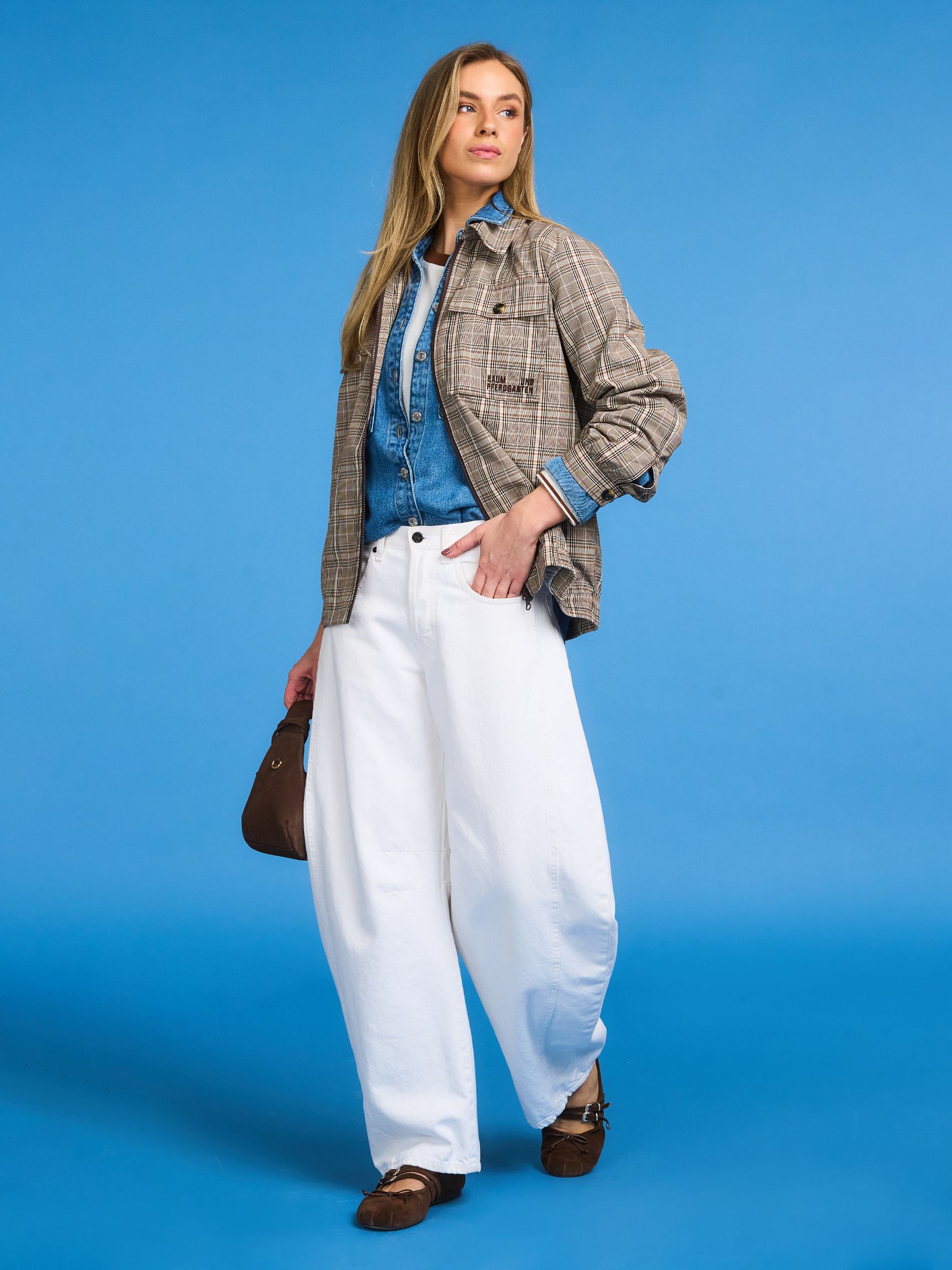 Woman in plaid jacket, denim shirt, and wide white pants against a blue background. She's holding a brown handbag and wearing brown shoes.