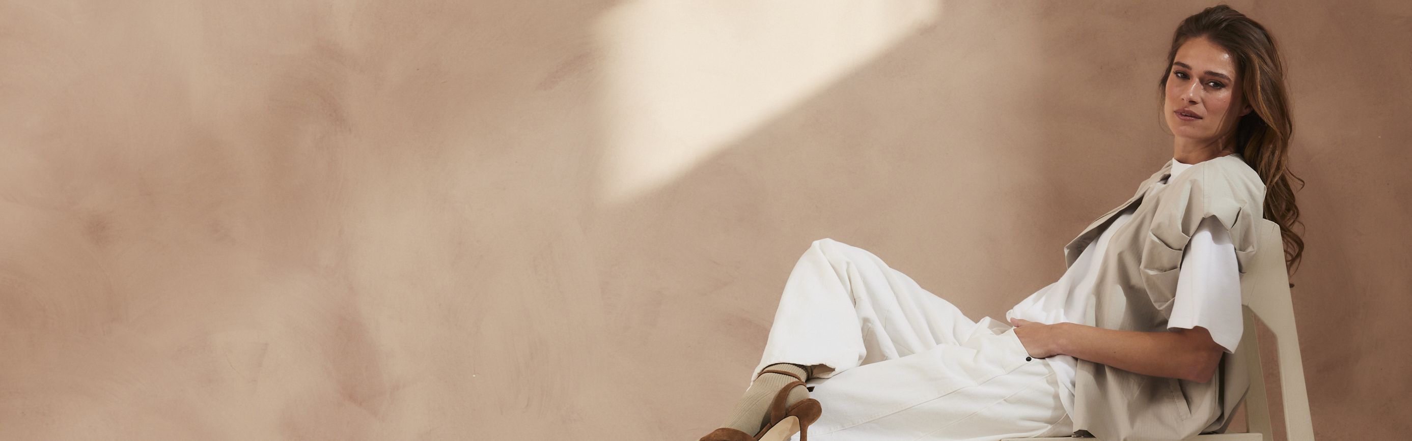 Woman in neutral-toned outfit sits relaxed on a chair against a soft beige background, with light softly illuminating the scene.